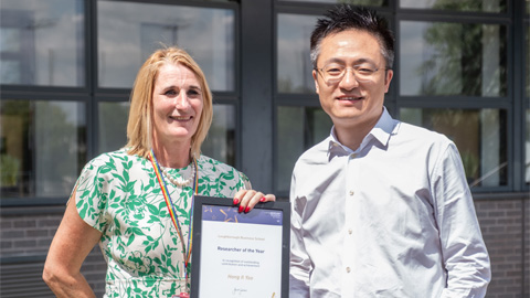 Two people standing in front of a building with large windows; one holding a framed certificate